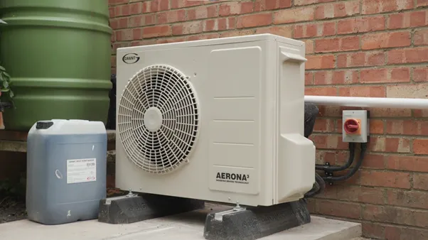 The installed air source heat pump on its mounts beside the brick wall at the rear of the house