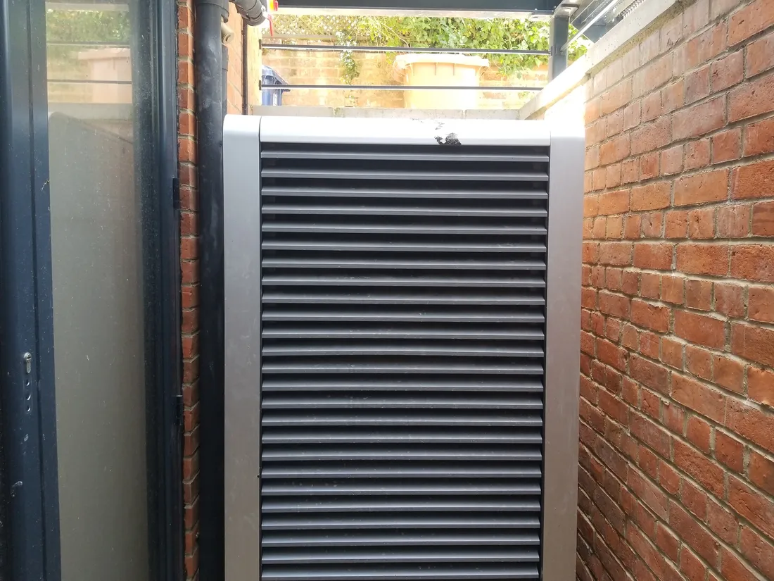 The external heat pump unit tucked into the narrow side passage between the brick walls